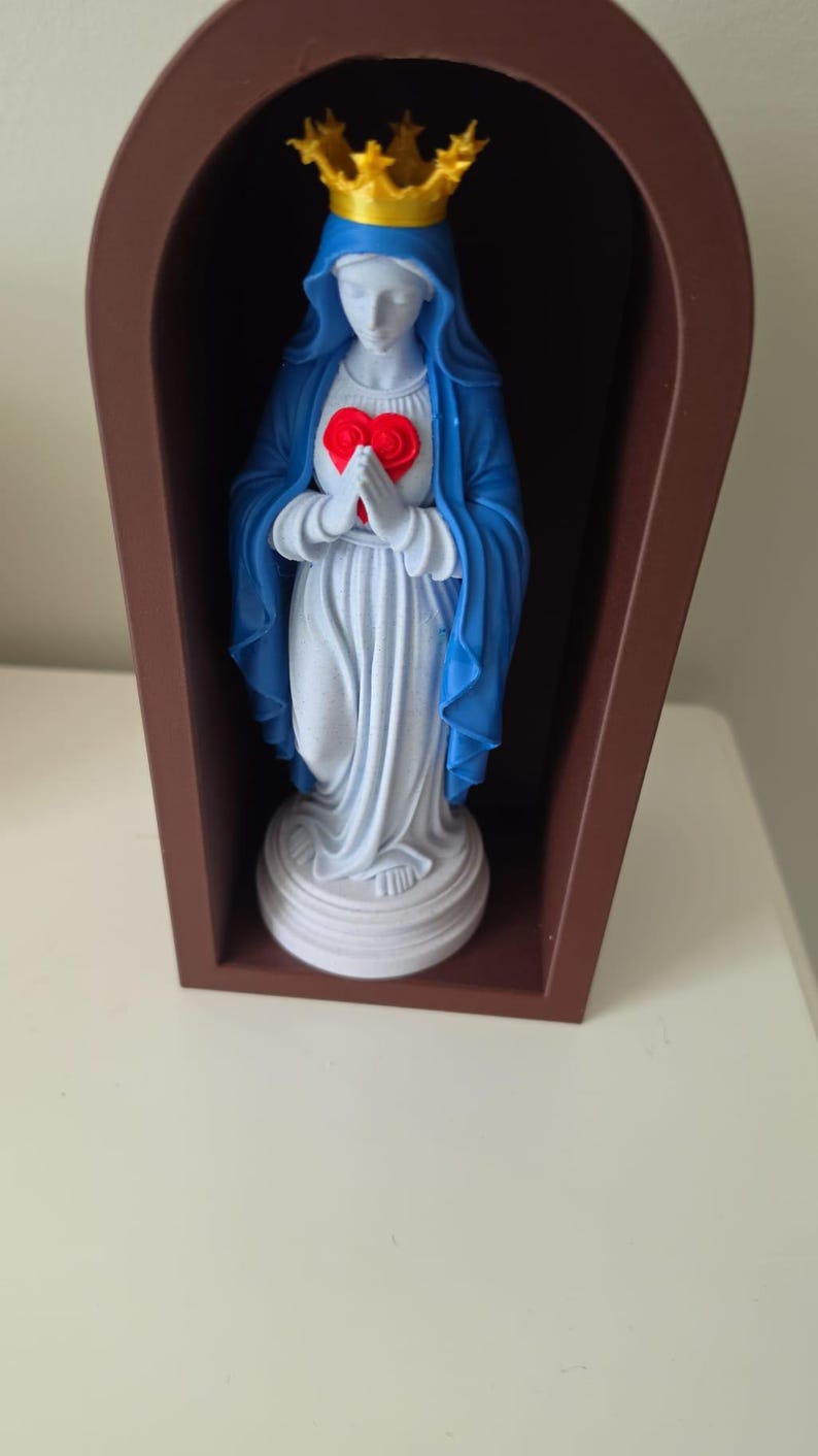 Puede incluir: Una figura blanca que viste una capa azul y una corona dorada, sosteniendo un coraz&oacute;n rojo con ambas manos. La figura est&aacute; colocada dentro de un marco marr&oacute;n arqueado de frente abierto sobre una base circular blanca.