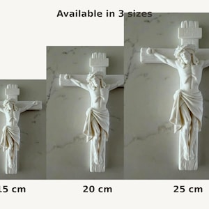May include: Three white wall crucifixes featuring a sculpted figure of Jesus on the cross. The text "Available in 3 sizes" appears above, with "15 cm", "20 cm", and "25 cm" labels below each respective cross, indicating the available height options.