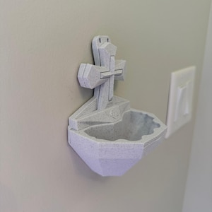 May include: A light gray, speckled, 3D-printed wall-mounted holy water font featuring a central cross design. The basin has a faceted, geometric shape with a small, raised emblem at the base of the cross, designed for attachment to a flat vertical surface.