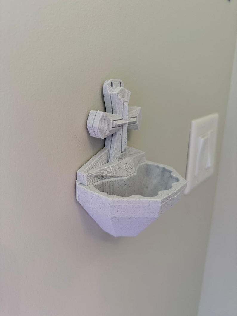 May include: A light gray, speckled, 3D-printed wall-mounted holy water font featuring a central cross design. The basin has a faceted, geometric shape with a small, raised emblem at the base of the cross, designed for attachment to a flat vertical surface.