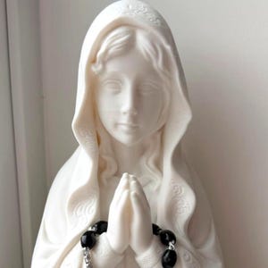 May include: A white matte statue of a figure with a draped head covering, depicted with hands pressed together in a prayer position. A rosary featuring black oval beads and a silver-toned metal chain is draped over the hands of the statue.