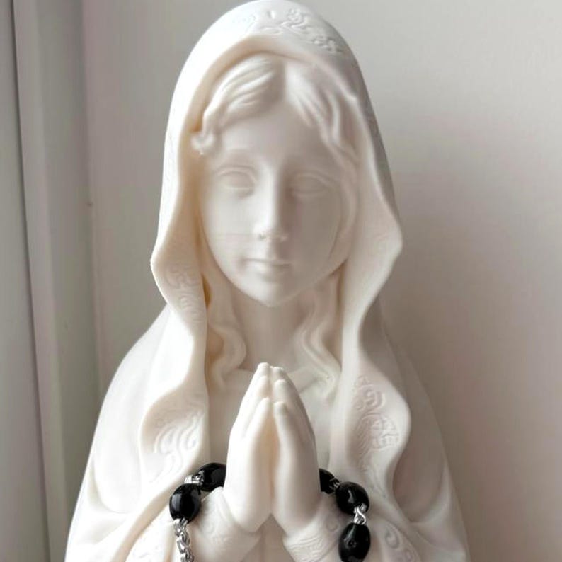 May include: A white matte statue of a figure with a draped head covering, depicted with hands pressed together in a prayer position. A rosary featuring black oval beads and a silver-toned metal chain is draped over the hands of the statue.