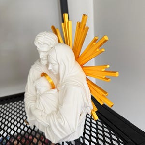 May include: A white matte figurine depicting a family of three figures huddled together. The figures have smooth, sculpted features. Behind the figures is a bright yellow, sunburst-shaped halo with radiating spikes, and the infant has a gold halo.