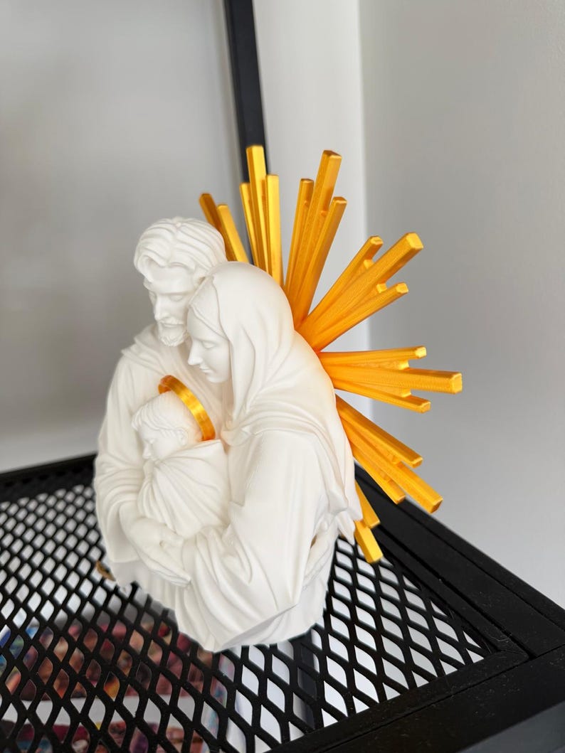 May include: A white matte figurine depicting a family of three figures huddled together. The figures have smooth, sculpted features. Behind the figures is a bright yellow, sunburst-shaped halo with radiating spikes, and the infant has a gold halo.