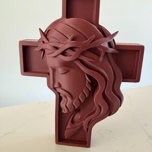 May include: A dark reddish-brown wall cross featuring a three-dimensional relief sculpture of a face wearing a crown of thorns. The sculpture is centred on the cross, with the head tilted downward and long, flowing hair depicted in layered segments.