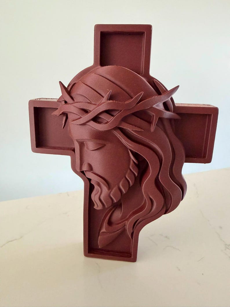 May include: A dark reddish-brown wall cross featuring a three-dimensional relief sculpture of a face wearing a crown of thorns. The sculpture is centred on the cross, with the head tilted downward and long, flowing hair depicted in layered segments.