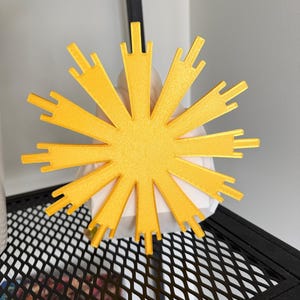 May include: A bright yellow, star-shaped decorative object with multiple radiating points of varying lengths. The object features a textured surface and is positioned in front of a white, geometric, faceted base, creating a high-contrast visual display.