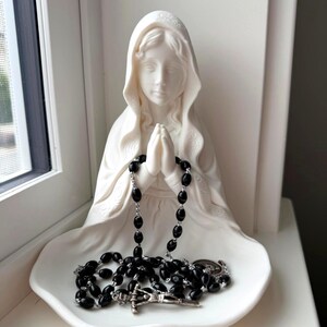 May include: A white matte ceramic figurine of a figure with a hooded head and hands clasped in prayer. A rosary with black oval beads and a silver-toned metal crucifix rests in the lap of the figurine, draped over the hands and the base.