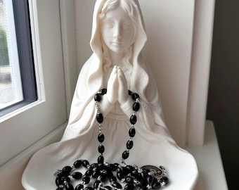 May include: A white matte ceramic figurine of a figure with a hooded head and hands clasped in prayer, featuring a wide base. A rosary with black oval beads and a silver-toned metal crucifix is draped over the hands and rests on the base.