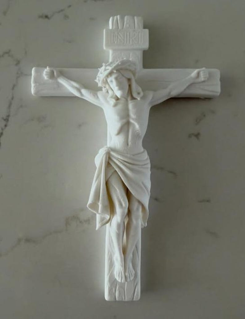May include: A white matte crucifix featuring a sculpted figure of a person wearing a crown of thorns and a draped cloth. The cross includes a top plaque with the embossed text INRI. The design is monochromatic with detailed relief work.