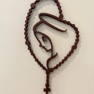 May include: A dark brown wall-mounted sculpture depicting a stylised profile of a figure with a halo, framed by a continuous loop of rosary beads that forms a heart shape, terminating at the bottom with a small cross pendant.
