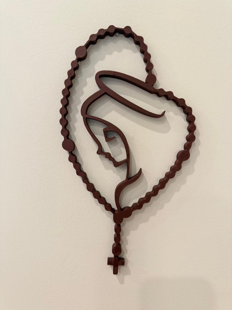 May include: A dark brown wall-mounted sculpture depicting a stylised profile of a figure with a halo, framed by a continuous loop of rosary beads that forms a heart shape, terminating at the bottom with a small cross pendant.