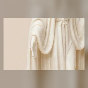 May include: A cream-colored statue depicting a figure draped in flowing, textured robes. The sculpture features a visible hand extended forward with fingers slightly parted, and the fabric of the garment shows intricate folds and a tied waist.