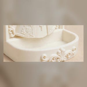 Puede incluir: Una pila de agua bendita de pared en color blanco roto, hecha de un material liso. El cuenco presenta un frente curvo con un dise&ntilde;o floral en relieve. Sobre el cuenco, una figura esculpida muestra detalles tallados y una cruz.
