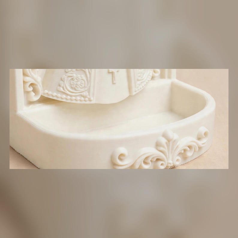 Puede incluir: Una pila de agua bendita de pared en color blanco roto, hecha de un material liso. El cuenco presenta un frente curvo con un dise&ntilde;o floral en relieve. Sobre el cuenco, una figura esculpida muestra detalles tallados y una cruz.
