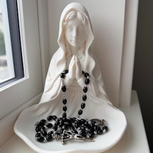 May include: A white matte figurine of a figure with a hooded head and hands clasped in prayer, featuring a wide, shallow base. A rosary with black oval beads and a silver-toned metal crucifix rests in the lap of the figurine on the base.