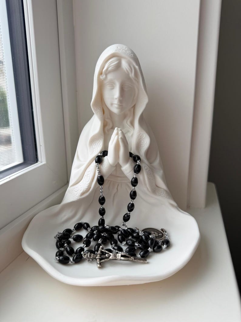 May include: A white matte figurine of a figure with a hooded head and hands clasped in prayer, featuring a wide, shallow base. A rosary with black oval beads and a silver-toned metal crucifix rests in the lap of the figurine on the base.