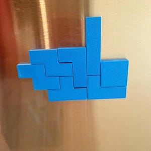 Op de afbeelding: Een set felblauwe geometrische kunststof blokken gerangschikt in een cluster op een metalen oppervlak. De stukken hebben de vorm van verschillende Tetris-achtige polyomino's, met een getextureerde matte afwerking en scherpe, strakke randen.