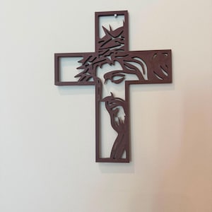 Puede incluir: Una cruz de pared de color marr&oacute;n oscuro que presenta la silueta recortada de un rostro con una corona de espinas. El dise&ntilde;o est&aacute; integrado en las barras verticales y horizontales de la cruz, creando una estructura hueca de l&iacute;neas limpias.