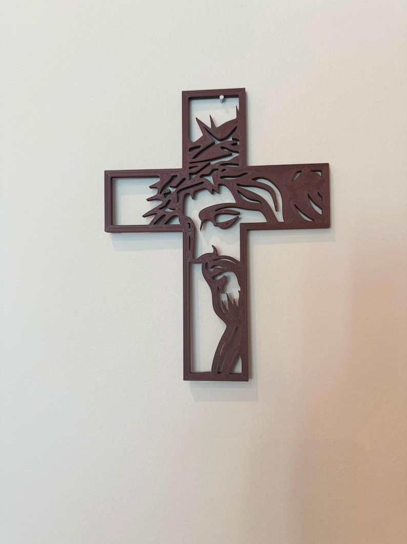 Puede incluir: Una cruz de pared de color marr&oacute;n oscuro que presenta la silueta recortada de un rostro con una corona de espinas. El dise&ntilde;o est&aacute; integrado en las barras verticales y horizontales de la cruz, creando una estructura hueca de l&iacute;neas limpias.