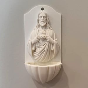 May include: A white wall-mounted holy water stoup featuring a relief sculpture of a bearded figure with long hair holding a heart at the chest. The piece has a semi-circular basin at the bottom and a small hole at the top for hanging on a wall.