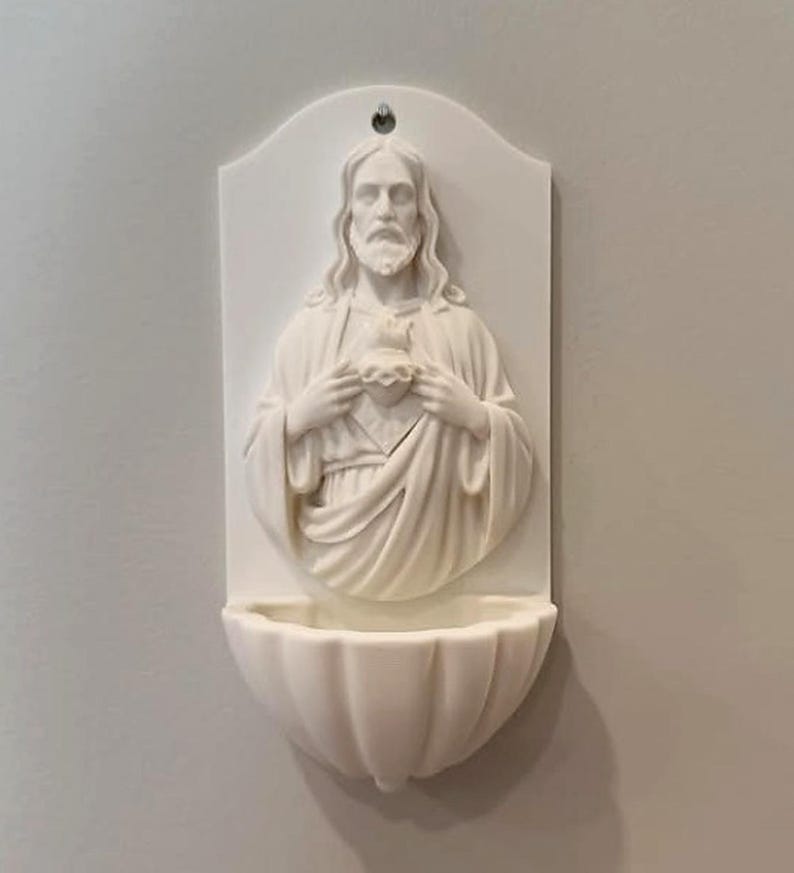 May include: A white wall-mounted holy water stoup featuring a relief sculpture of a bearded figure with long hair holding a heart at the chest. The piece has a semi-circular basin at the bottom and a small hole at the top for hanging on a wall.