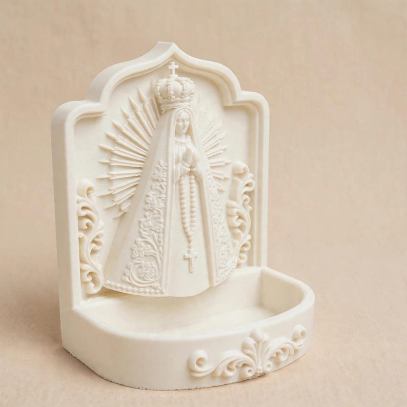 Puede incluir: Una pila de agua bendita de pared en color crema con un relieve de la Virgen Mar&iacute;a que lleva una corona y sostiene un rosario. La figura est&aacute; rodeada por un halo de rayos y adornos de volutas, con un cuenco en la parte inferior.