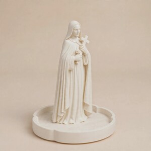 May include: A cream-colored figurine of a figure wearing a hooded robe, holding a small cross and a rose in their hands. The statue stands on a circular, scalloped-edge base, featuring a smooth, matte finish throughout the entire structure.