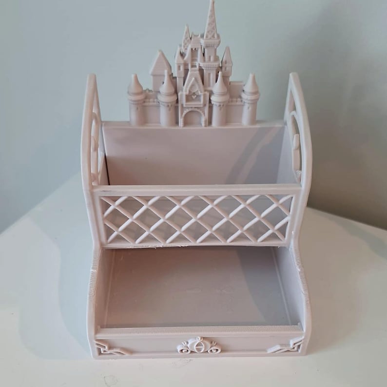 May include: A light grey plastic desk organiser featuring a castle silhouette at the top, a lattice-patterned middle section, and a small carriage emblem centred on the bottom front edge. The structure has two open compartments for storage.