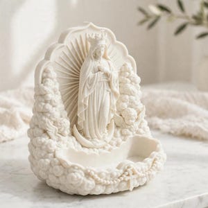 May include: A cream-colored resin sculpture featuring a central figure of the Virgin Mary wearing a crown and standing on a crescent moon. The figure is surrounded by clouds, cherubs, and a sunburst background, with a small basin at the base.