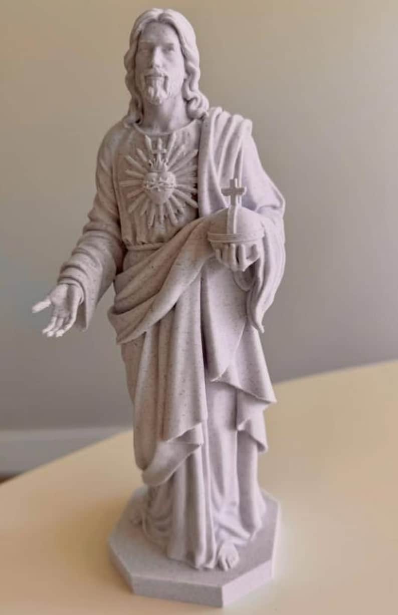 May include: A light gray speckled statue of a figure with long hair and a beard, wearing draped robes. The figure has a Sacred Heart emblem on the chest, holds a globus cruciger in one hand, and has the other hand extended forward.