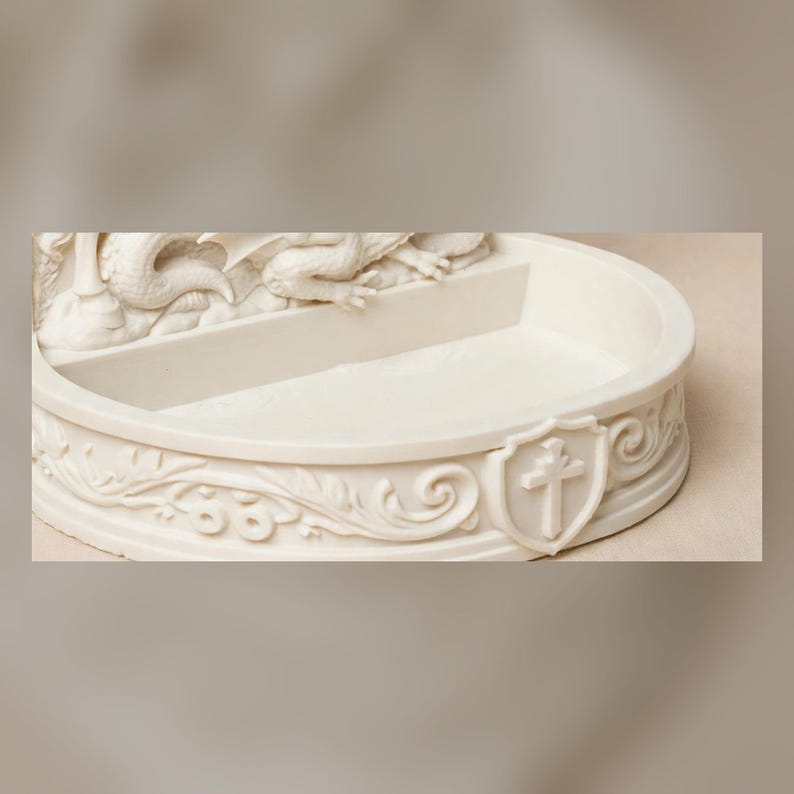 K&ouml;nnte beinhalten: Eine cremefarbene, halbkreisf&ouml;rmige Steinschale mit einer skulptierten Drachenfigur am hinteren Rand. Die Vorderseite ist mit gepr&auml;gten Schn&ouml;rkeln und einem zentralen Wappenschild verziert, das ein erhabenes Kreuzsymbol zeigt.