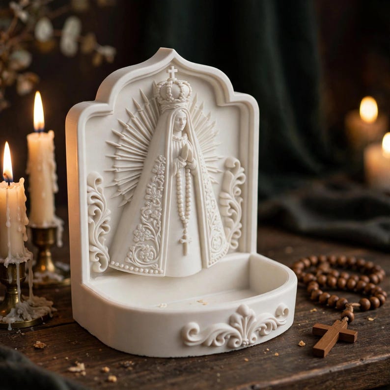 Puede incluir: Una pila de agua bendita de color blanco, hueso o crema que presenta una escultura en relieve de la Virgen Mar&iacute;a con corona y un rosario. El panel trasero tiene un dise&ntilde;o de rayos de sol y la base es un cuenco poco profundo con volutas.