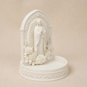 May include: An off-white resin sculpture featuring a central figure of the Virgin Mary with hands clasped in prayer. The figure is framed by an arched backdrop with floral rose carvings and a circular base with decorative scrollwork detailing.