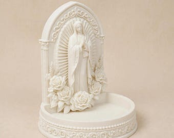 May include: An off-white resin sculpture featuring a central figure of the Virgin Mary with hands clasped in prayer. The figure is framed by an arched backdrop with floral rose carvings and a circular base with decorative scrollwork detailing.
