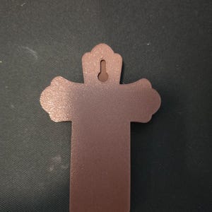 May include: A solid brown cross with a textured surface and a keyhole-shaped mounting slot at the top. The cross features rounded, scalloped edges on the top and side arms, and a flat, straight-edged base, designed for wall mounting.