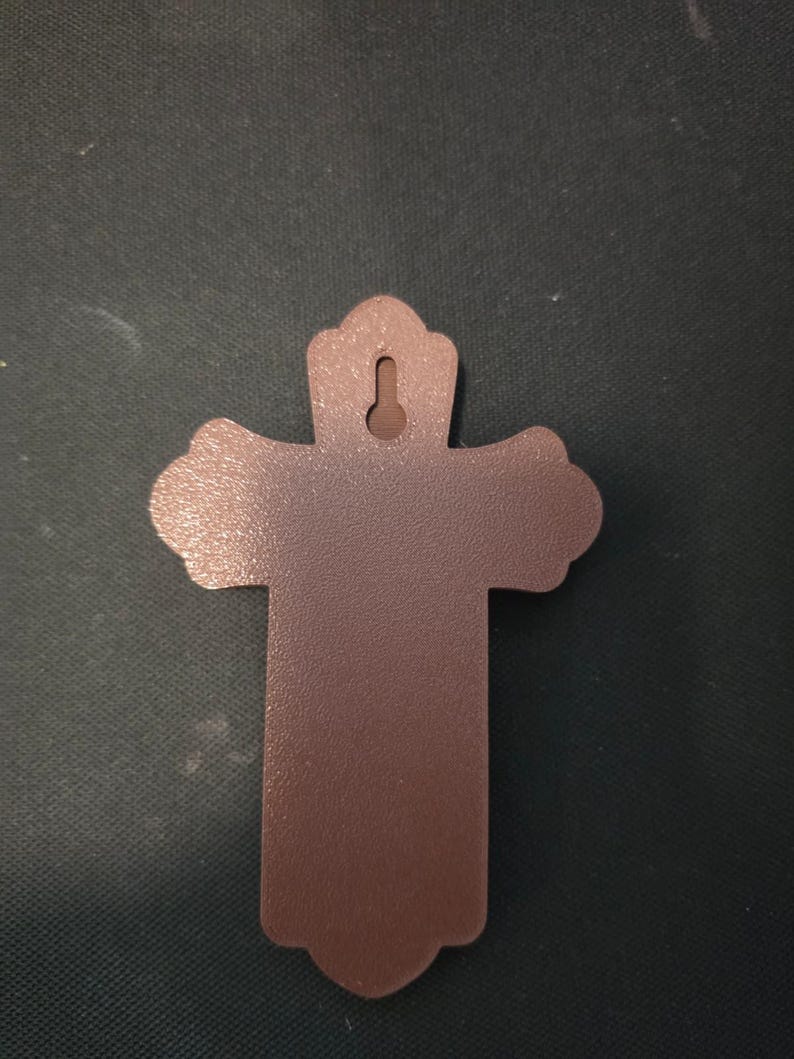 May include: A solid brown cross with a textured surface and a keyhole-shaped mounting slot at the top. The cross features rounded, scalloped edges on the top and side arms, and a flat, straight-edged base, designed for wall mounting.