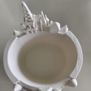 Pu&ograve; includere: Un contenitore cilindrico bianco stampato in 3D con una struttura a castello in miniatura integrata nel bordo superiore. La parete esterna presenta piccole sporgenze a forma di torretta, mentre la base interna mostra motivi circolari concentrici.