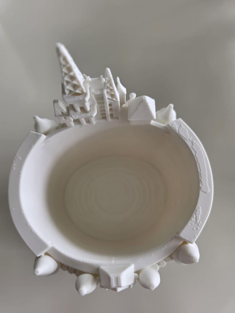 Pu&ograve; includere: Un contenitore cilindrico bianco stampato in 3D con una struttura a castello in miniatura integrata nel bordo superiore. La parete esterna presenta piccole sporgenze a forma di torretta, mentre la base interna mostra motivi circolari concentrici.
