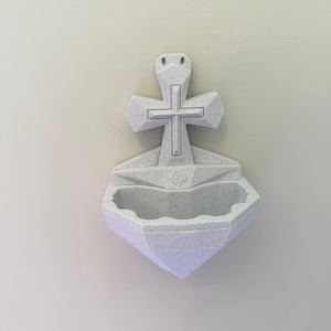 May include: A wall-mounted holy water font made of off-white speckled material. The design features a central cross positioned above a basin with a scalloped edge. The surface displays visible 3D printed layer lines throughout the entire structure.