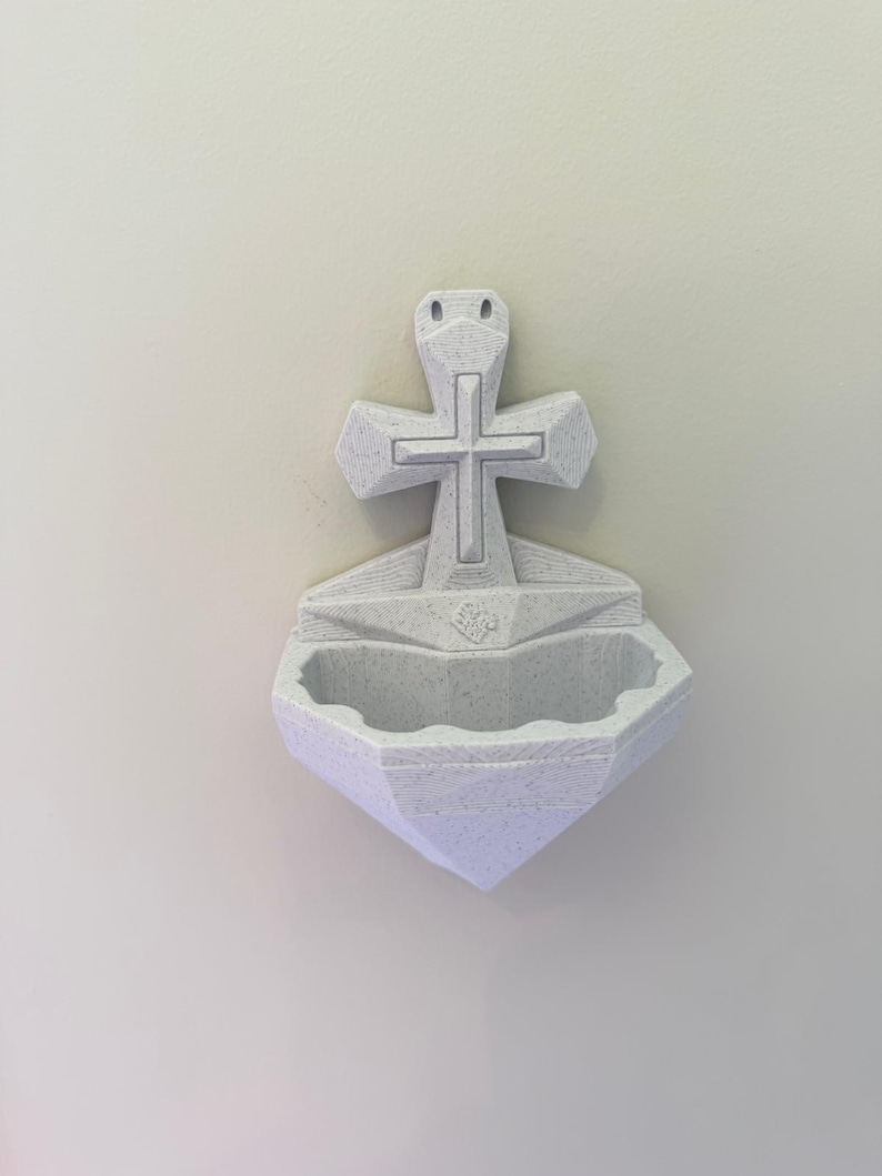 May include: A wall-mounted holy water font made of off-white speckled material. The design features a central cross positioned above a basin with a scalloped edge. The surface displays visible 3D printed layer lines throughout the entire structure.