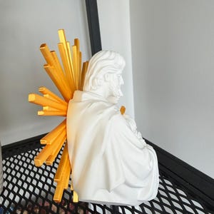 May include: A white bust of a bearded figure draped in a robe, viewed from the side. Behind the figure is a bright gold sunburst halo consisting of multiple radiating rectangular beams of varying lengths, extending outward from the center.