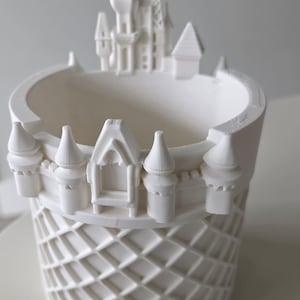 Pu&ograve; includere: Un vaso cilindrico bianco caratterizzato da un motivo decorativo a castello lungo il bordo superiore. La parete esterna del vaso presenta un motivo a reticolo in rilievo, mentre il bordo superiore &egrave; decorato con torri, torrette e un portale.