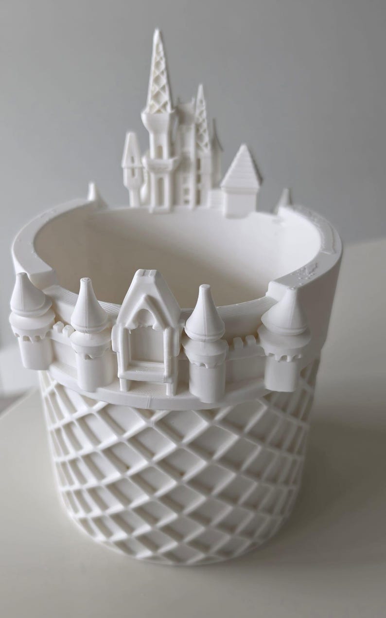 Pu&ograve; includere: Un vaso cilindrico bianco caratterizzato da un motivo decorativo a castello lungo il bordo superiore. La parete esterna del vaso presenta un motivo a reticolo in rilievo, mentre il bordo superiore &egrave; decorato con torri, torrette e un portale.