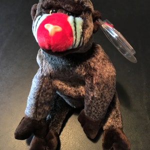 May include: A plush toy mandrill monkey with brown and gray fur, a red nose, and blue stripes on its face. The toy has dark brown feet and hands, and a clear tag is attached to its ear. The toy is sitting on a dark surface.