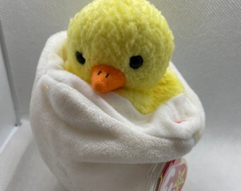 May include: A plush yellow chick toy with a soft, textured body, orange beak, and black eyes, emerging from a white egg. The egg is also plush and has a smooth texture. A tag is attached to the egg.