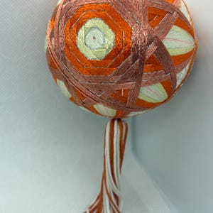May include: A traditional Japanese hand-stitched ornament called a "Temari" with a geometric pattern in shades of orange and white. The ornament has a long tassel made of orange and white threads.