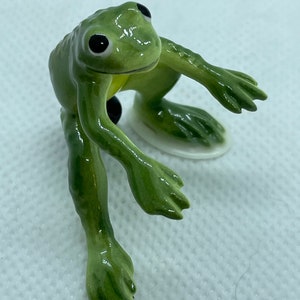 May include: A green ceramic frog figurine with black eyes. The frog is sitting on its hind legs with its front legs outstretched.