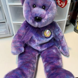 May include: Purple and blue tie-dye Beanie Baby bear with a purple ribbon around its neck and a Ty Beanie Babies Official Club tag. The bear has black eyes and a black nose.