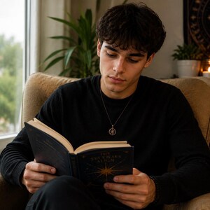 A young man in a black long-sleeve shirt sits in an armchair by a window reading an open book.